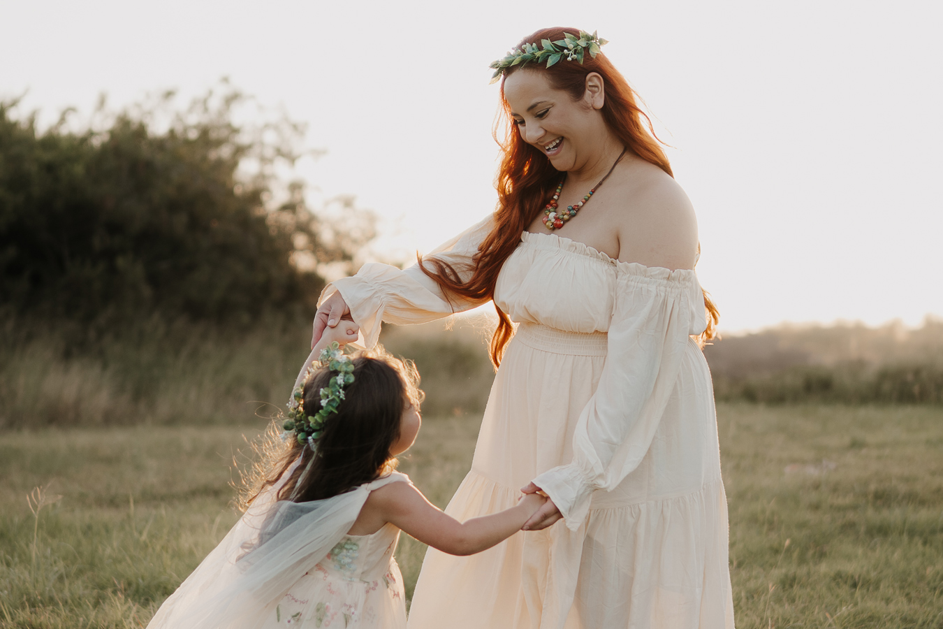 A woman with long red hair and a green leafy crown, wearing an off-the-shoulder cream-colored dress, smiles down at a young girl in a floral dress and matching crown as they hold hands in a grassy field. The warm light of the setting sun illuminates the scene. Photo by Valerie Marie Photography
