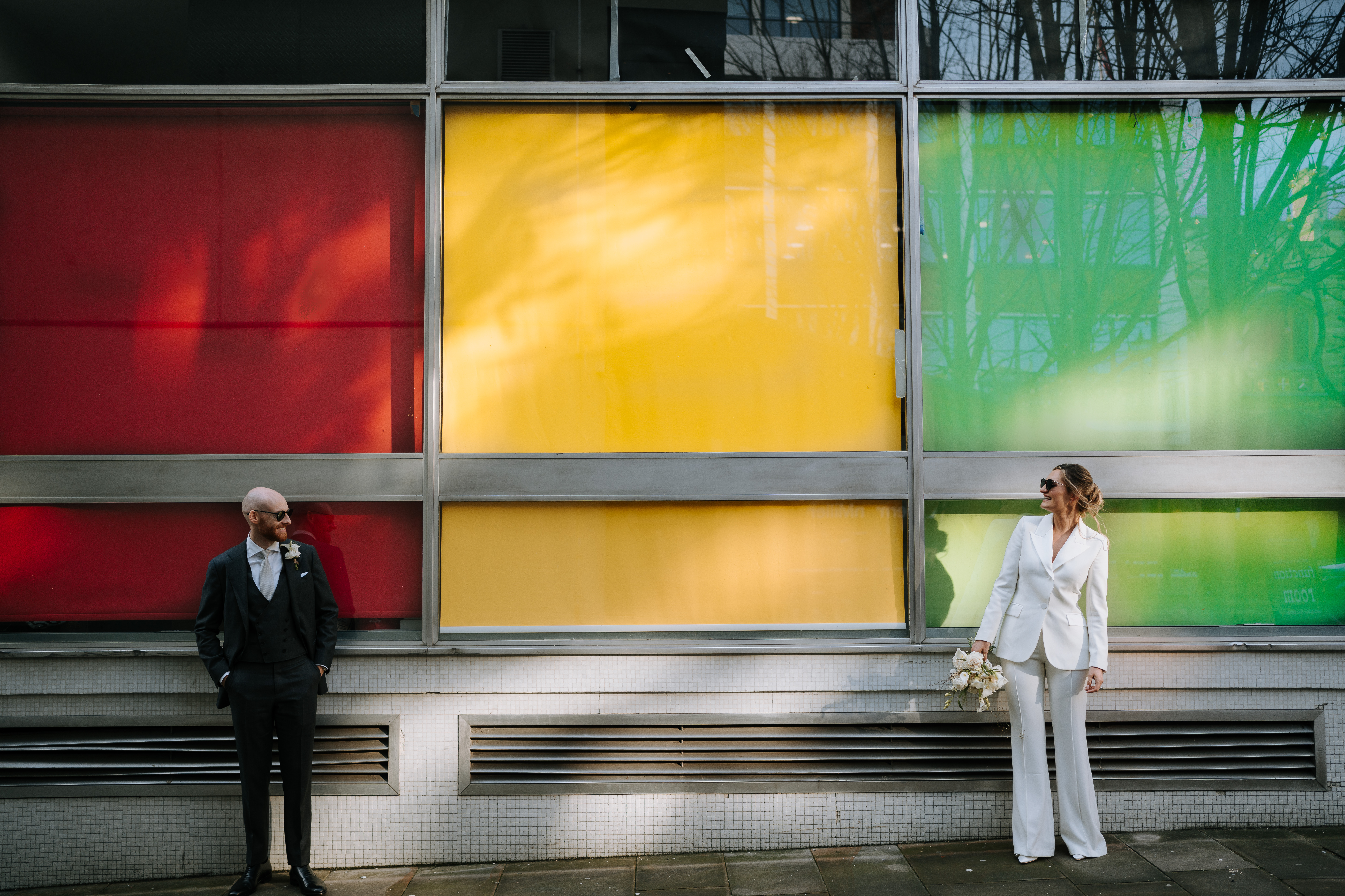 Wedding couple wearing suits in front of a colorful building. photo by Steve Bridgwood